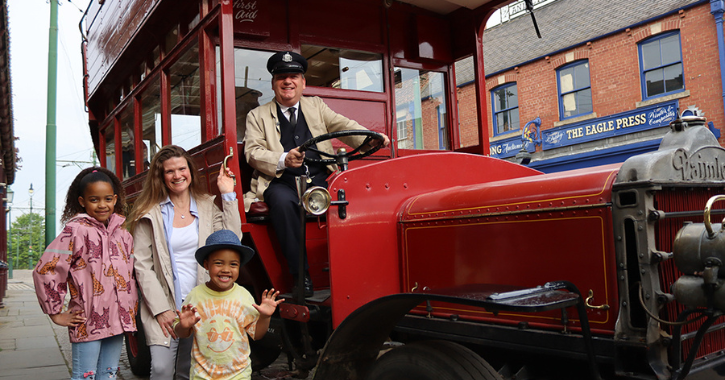 Mother with two children stand smiling next to a man in an historic red vehicle at Beamish Museum